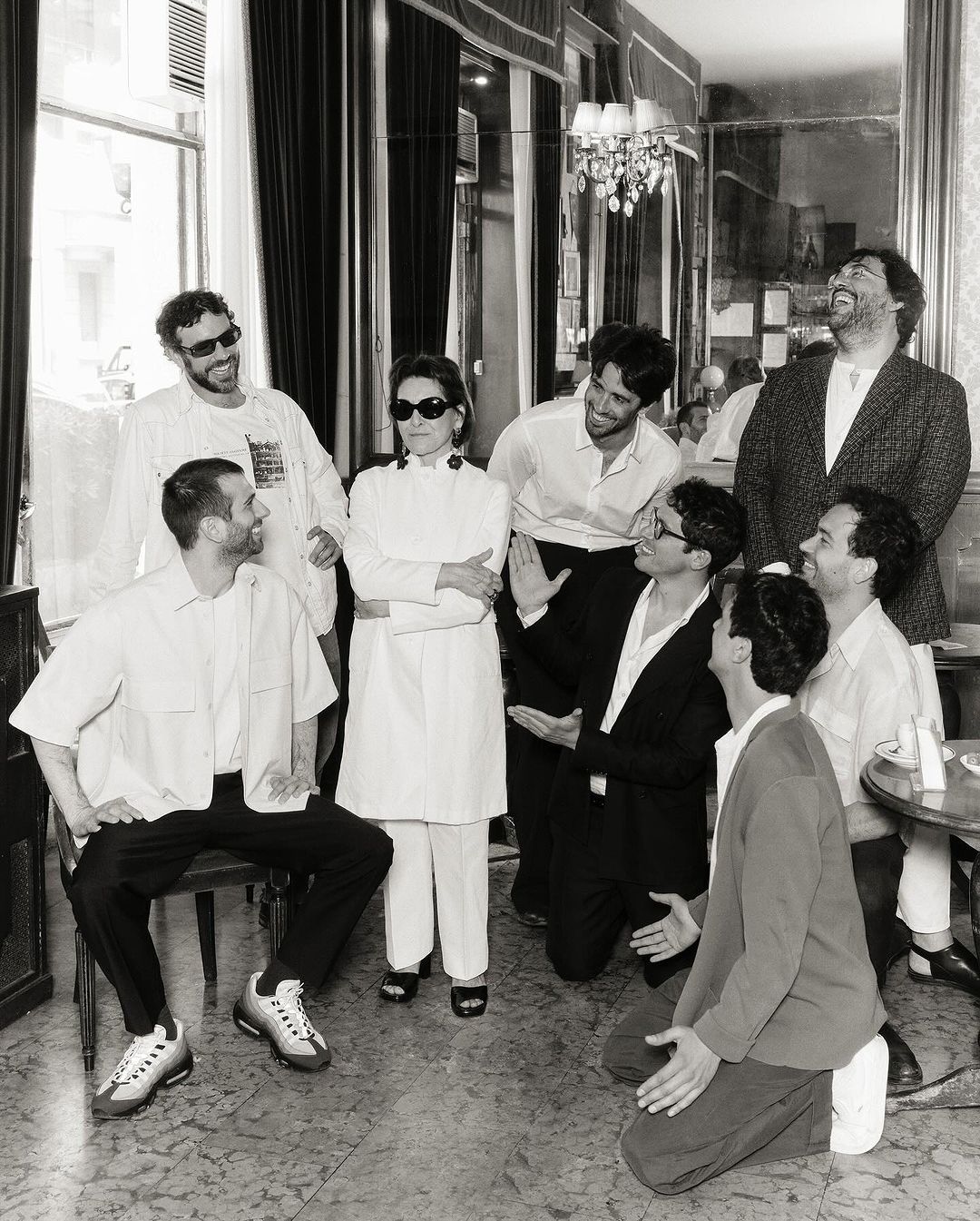 Black-and-white portrait of eight adults in a casual setting, with a chandelier and window visible in the background.