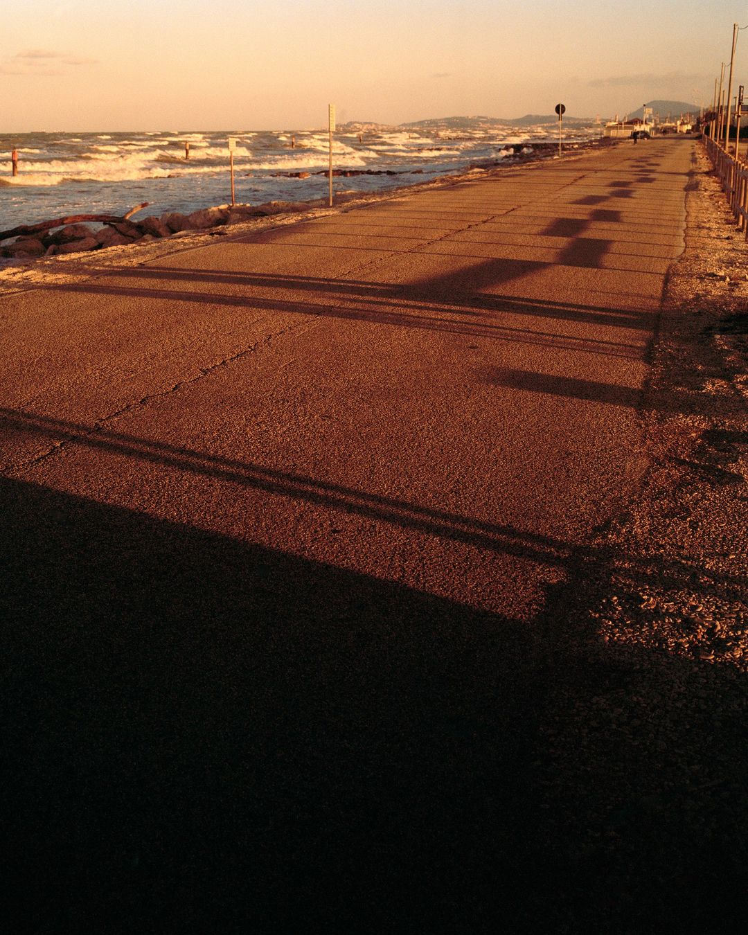 Seaside road with dark shadows, white waves crashing on the shore under a golden light. Distant buildings are visible along the coastline.