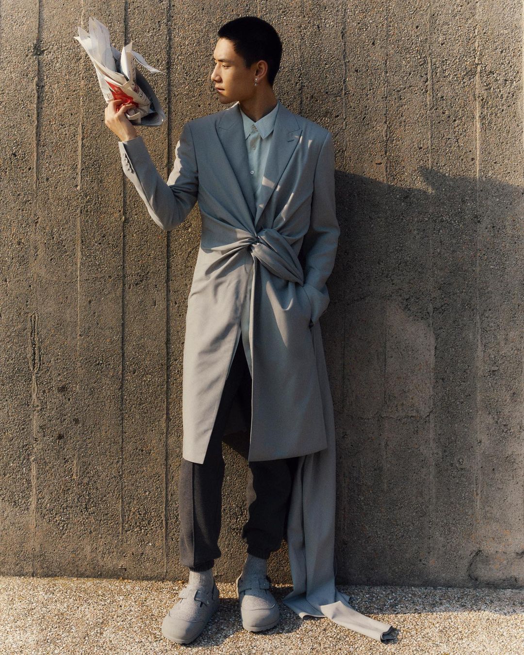 Asian man in a gray suit holds a newspaper. The man wears dark pants and gray shoes while posing against a concrete wall outdoors.