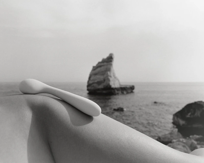 Monochrome shot of a person's arm with a white implement against a sea backdrop featuring a rocky island and overcast sky.