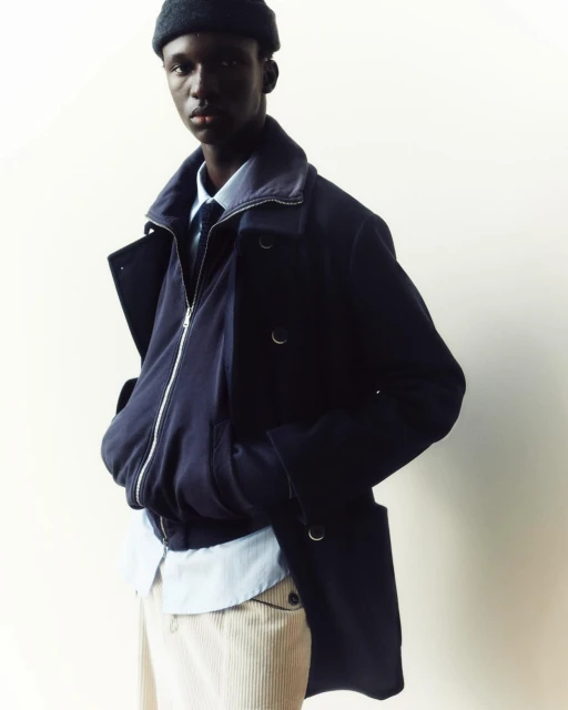 An African American male model poses in a navy blue overcoat, blue bomber jacket, baby-blue dress shirt, and corduroy pants against an off-white backdrop.