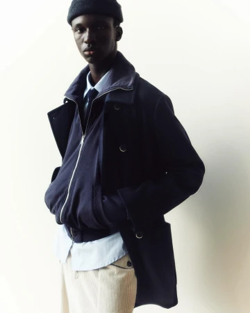 An African American male model poses in a navy blue overcoat, blue bomber jacket, baby-blue dress shirt, and corduroy pants against an off-white backdrop.