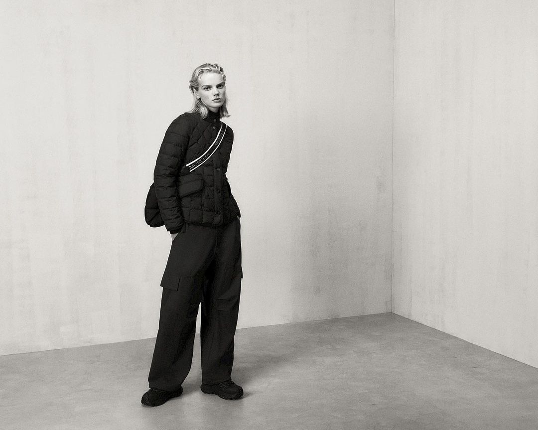 Fashion shot in a photo studio features a model wearing a black puffer jacket and loose pants with gray wall and floor in the background.