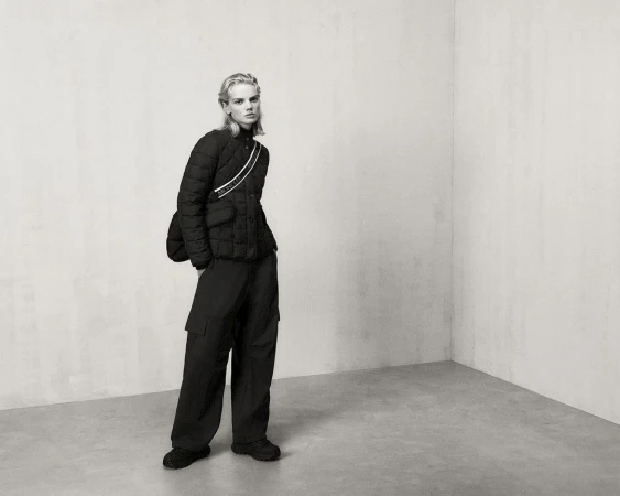 Fashion shot in a photo studio features a model wearing a black puffer jacket and loose pants with gray wall and floor in the background.