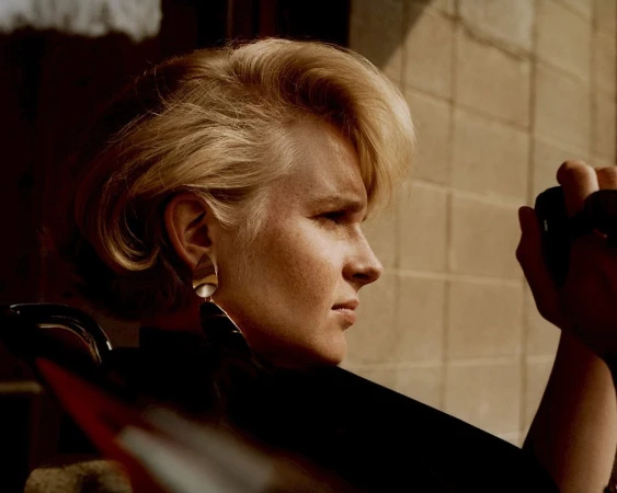 Profile portrait of a blonde woman wearing statement earrings against a concrete block wall background