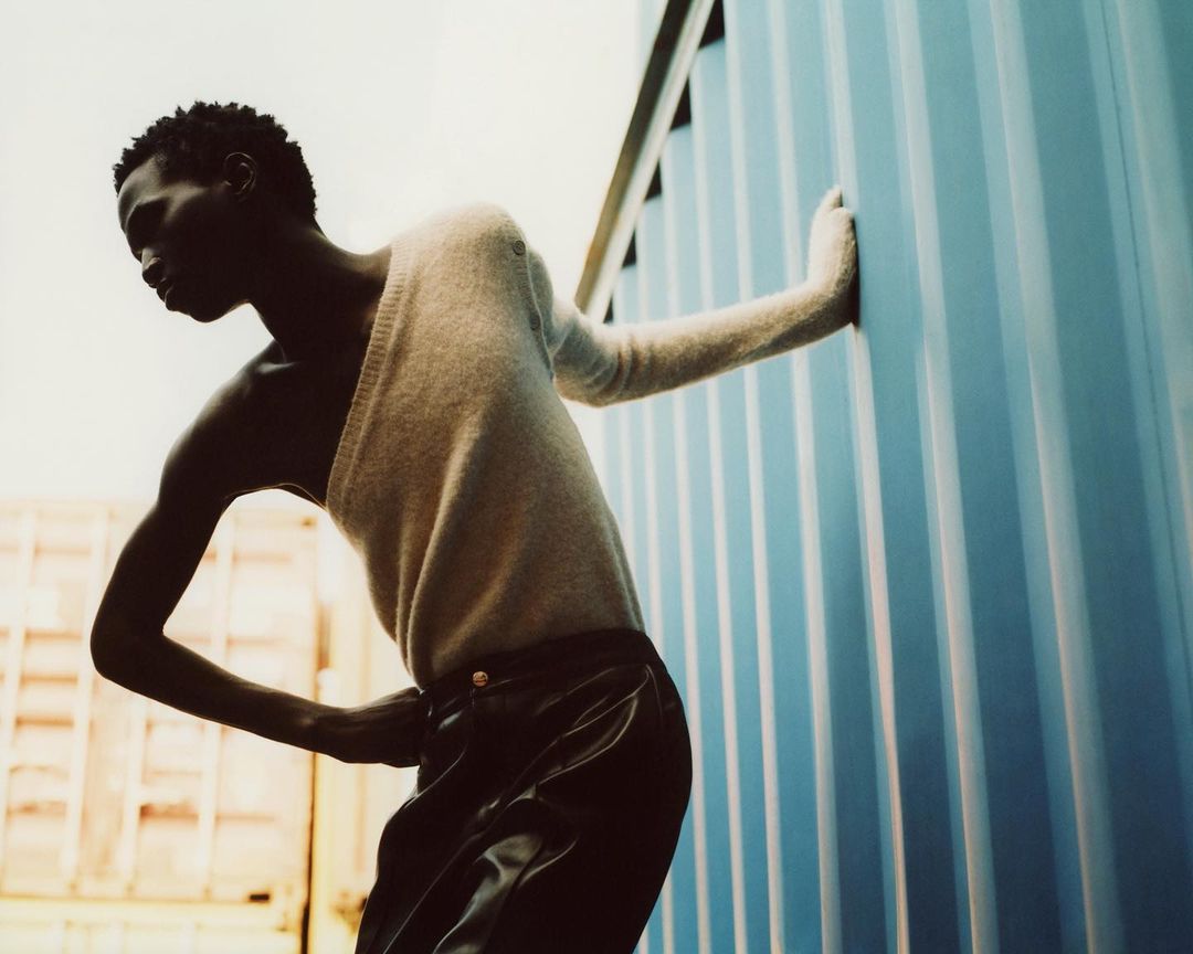 Athletic, dark-skinned male model poses in asymmetric sweater and leather pants, leaning against a blue corrugated metal wall in outdoor setting.