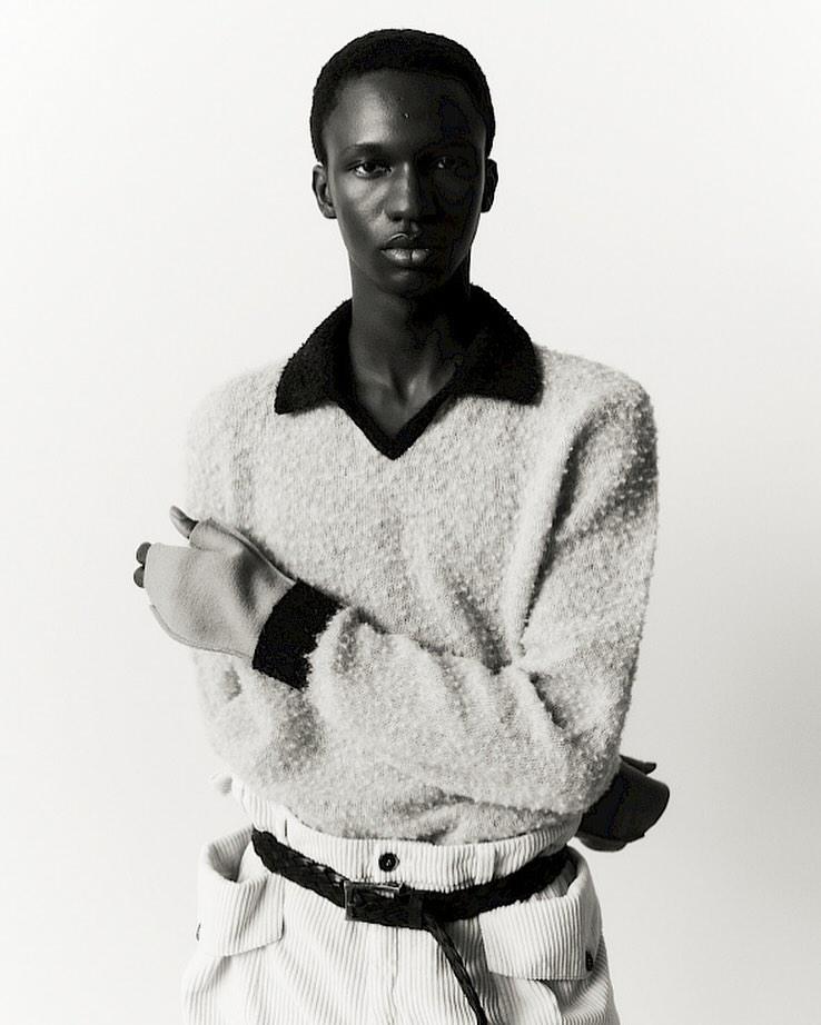 Portrait of a Black male model in a fuzzy sweater and corduroy pants against a bright white backdrop in a studio setting.