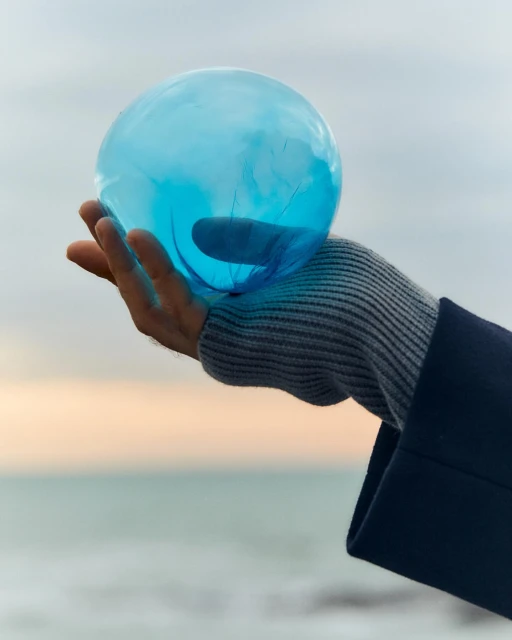 Close-up of a hand holding a translucent blue glass orb against a soft, blurred background of sky and ocean, creating a serene, ethereal scene.