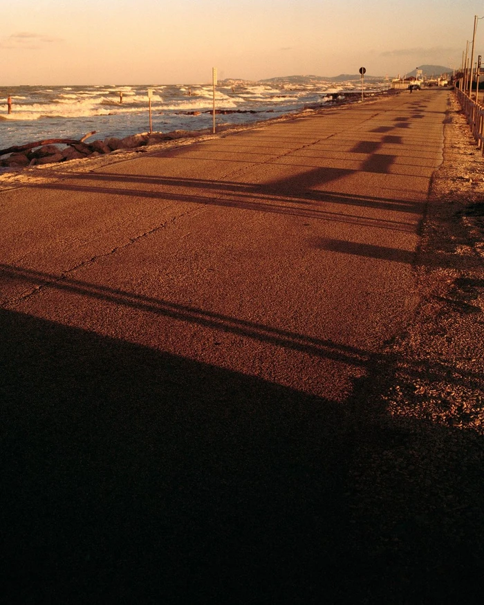 A paved coastal road beside a choppy sea at sunset, casting long dark shadows across the asphalt.