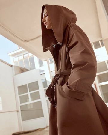 A woman wearing a brown hooded coat with a tied belt stands outdoors against a light building exterior.
