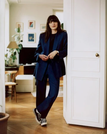Woman in a blue blazer and matching trousers standing and leaning against a white doorway in an apartment interior.