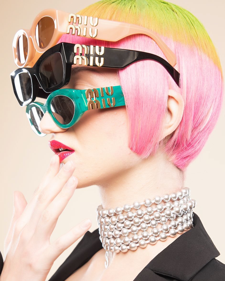Close-up of a fair-skinned woman with vibrant pink and green hair wearing three pairs of Miu Miu sunglasses and a pearl choker.