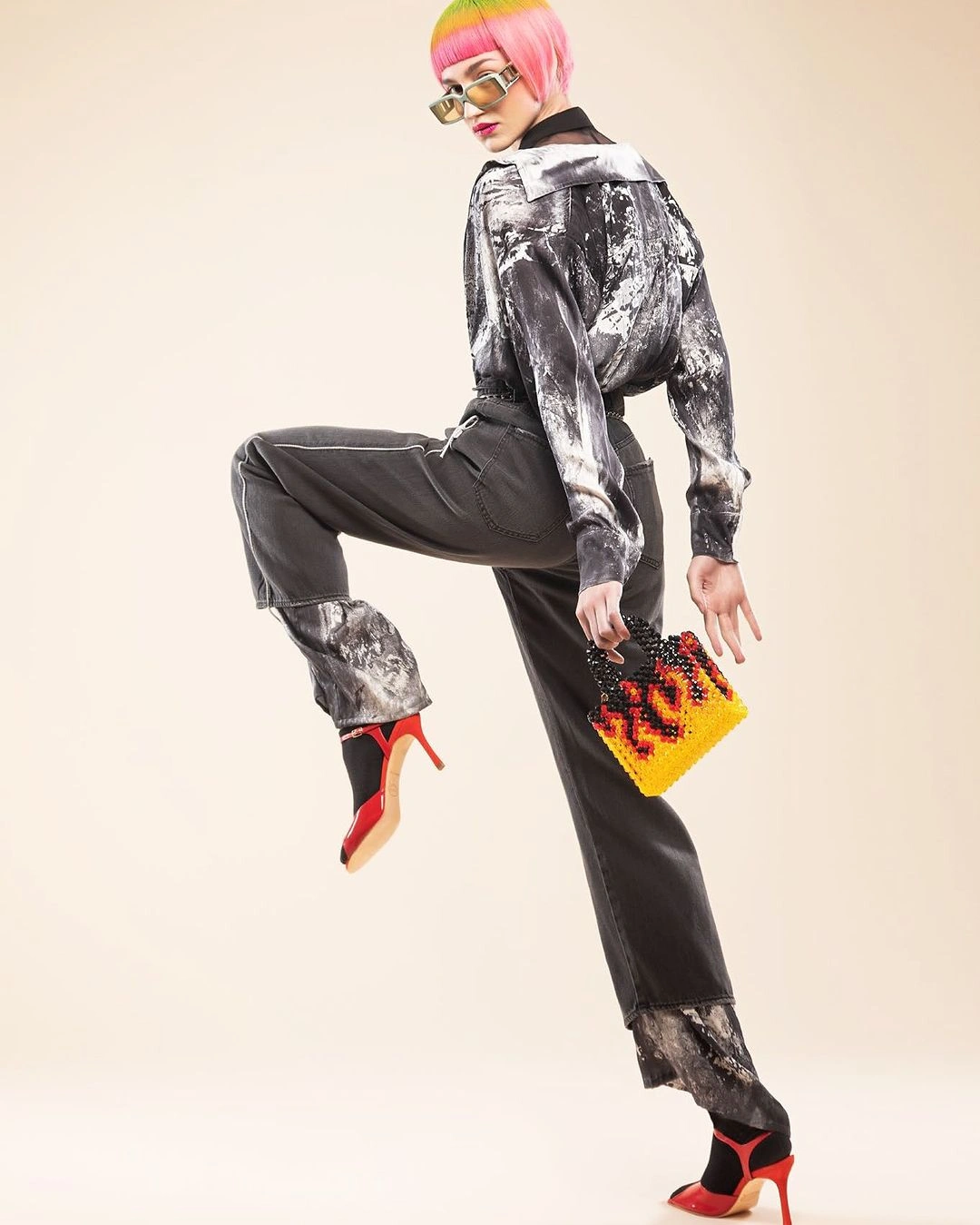Model with pink hair and sunglasses wearing a printed shirt, dark trousers, and red heels against a plain background
