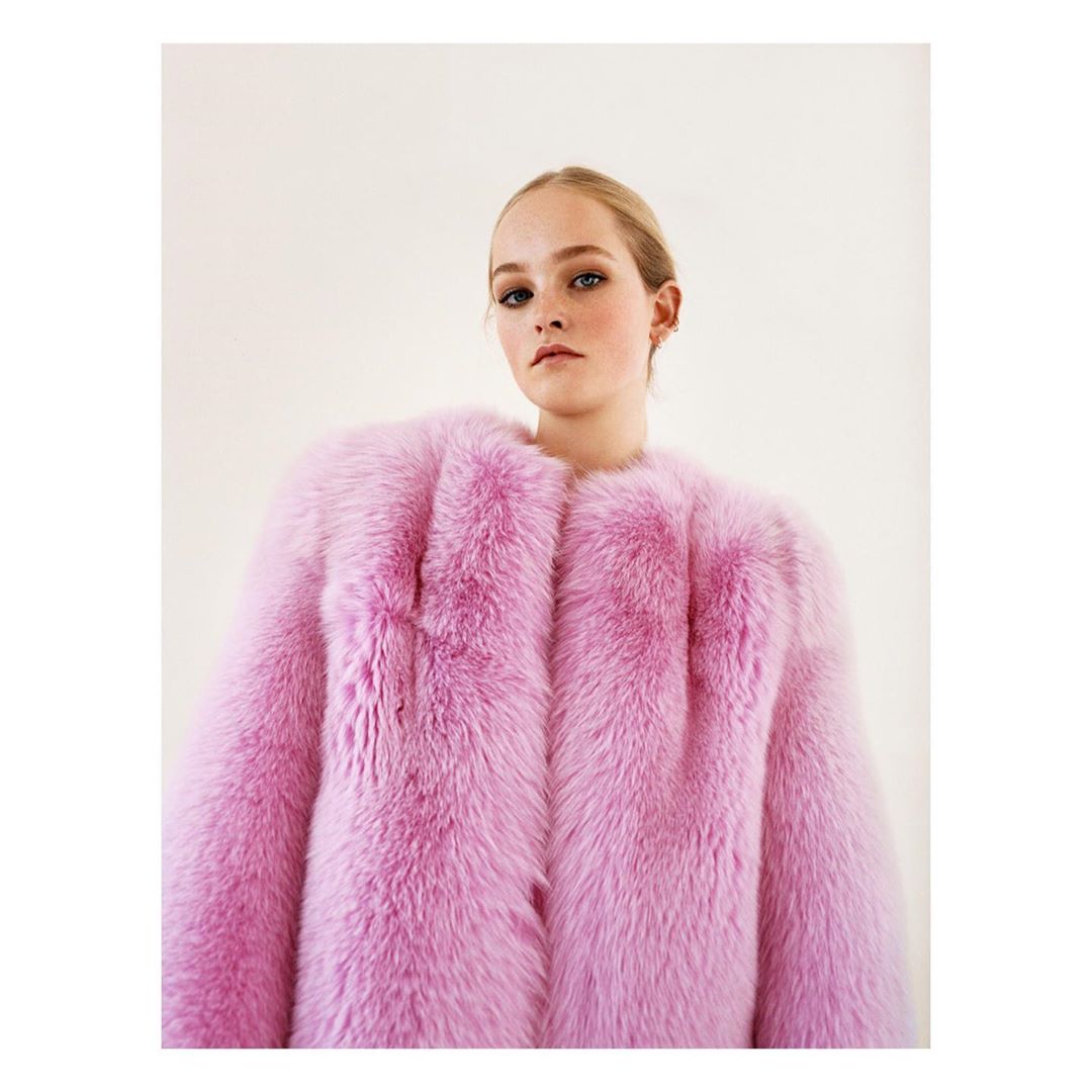 Fashion portrait of a young woman wearing a bright pink fur coat, posing indoors in front of a neutral background, lit with soft lighting.