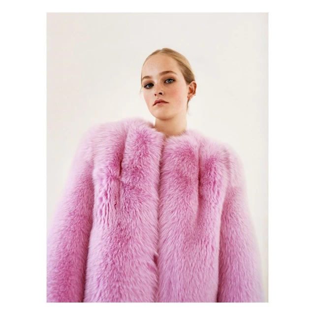 Fashion portrait of a young woman wearing a bright pink fur coat, posing indoors in front of a neutral background, lit with soft lighting.