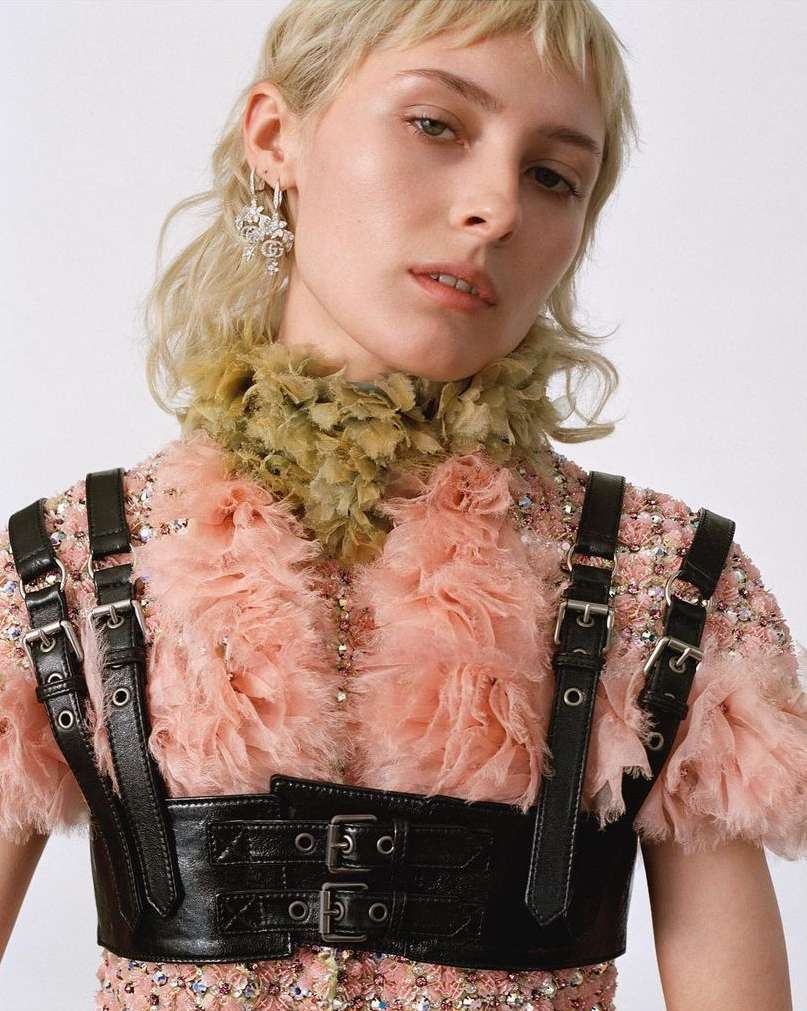 Close-up of a blonde woman wearing a pink ruffled dress and a black harness, with a white background in a fashion studio setting.