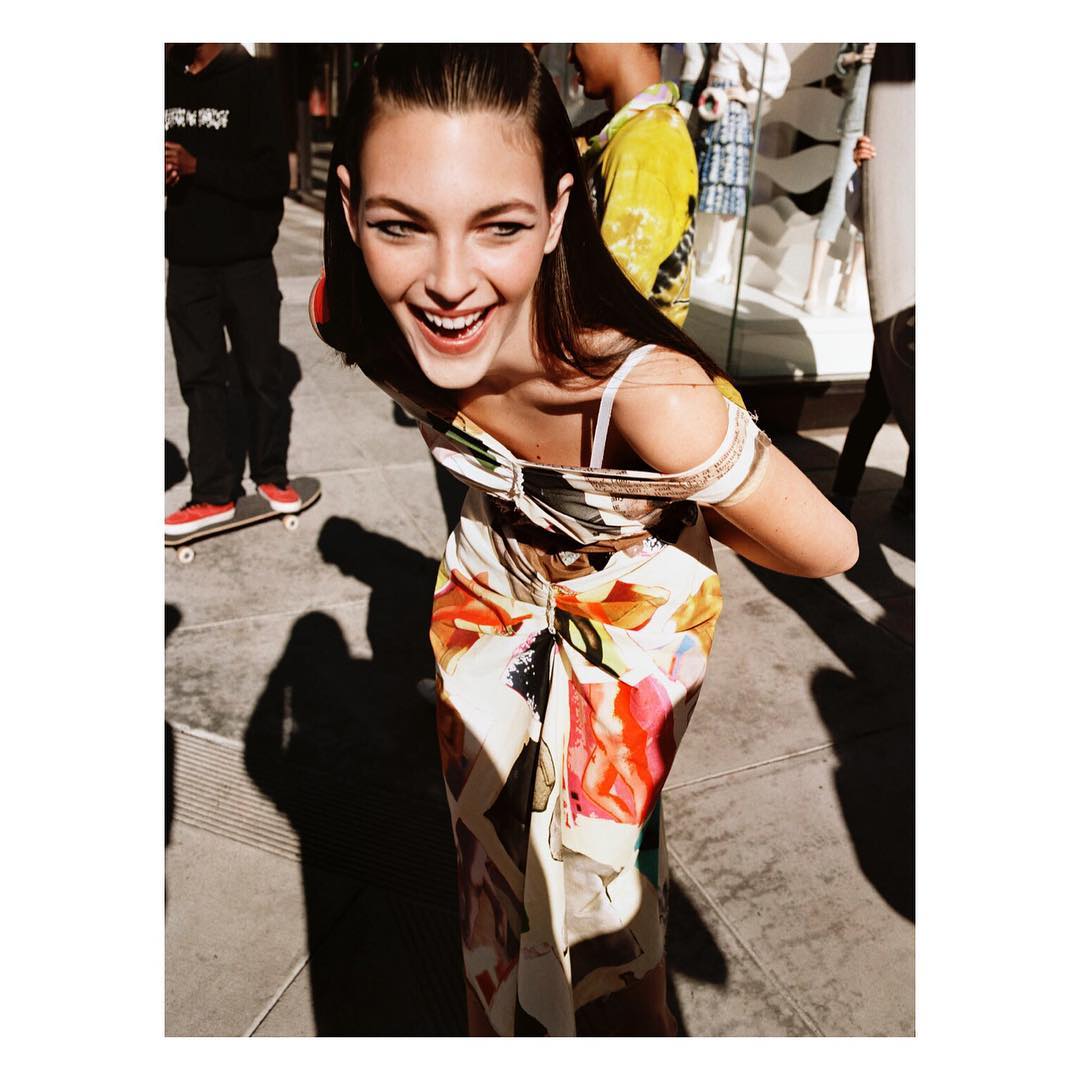 Smiling woman in floral dress on city sidewalk with passersby. Casual fashion shot with natural light and urban setting.