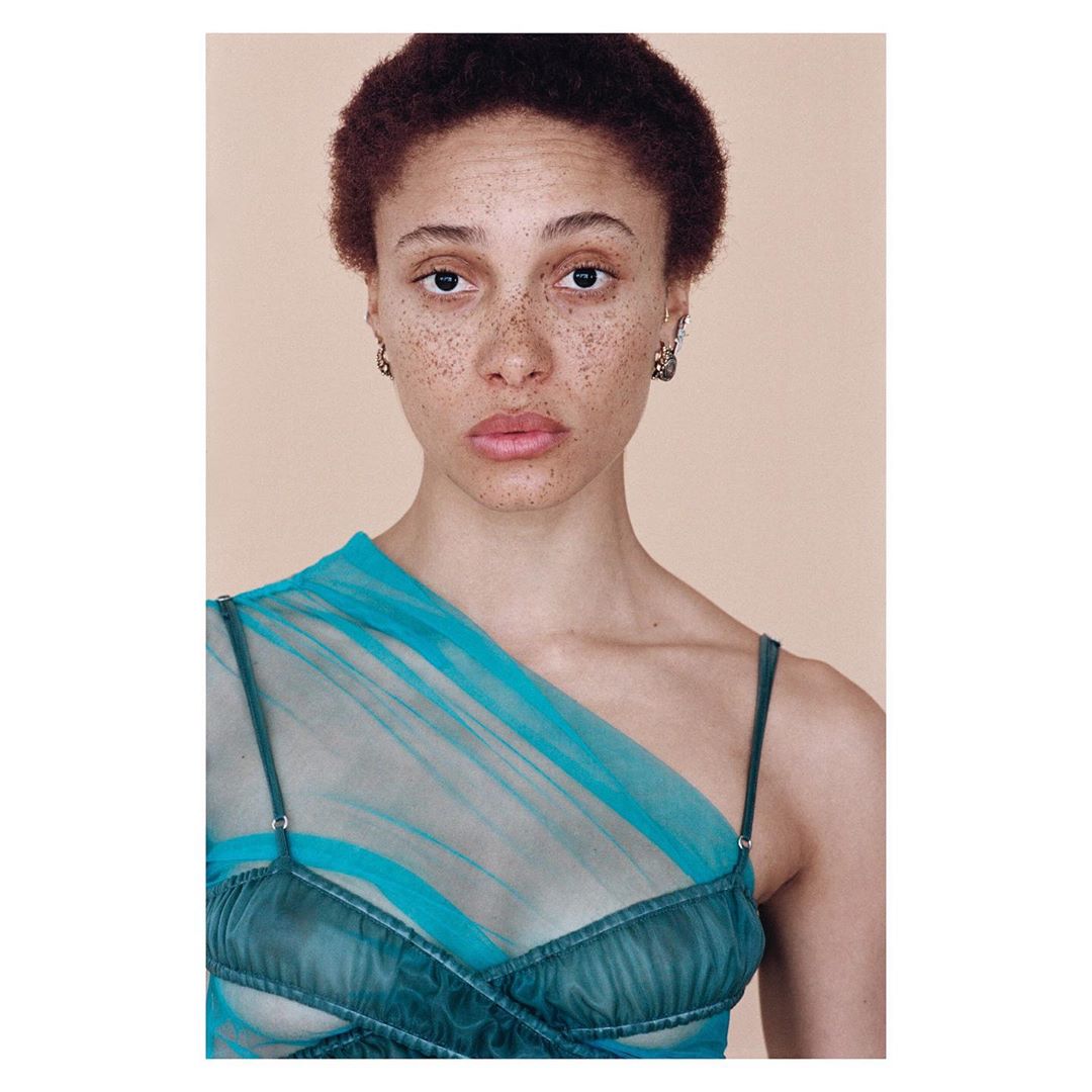 Portrait of a freckled adult female with short curly auburn hair, wearing a semi-sheer teal dress against a peach-colored backdrop.