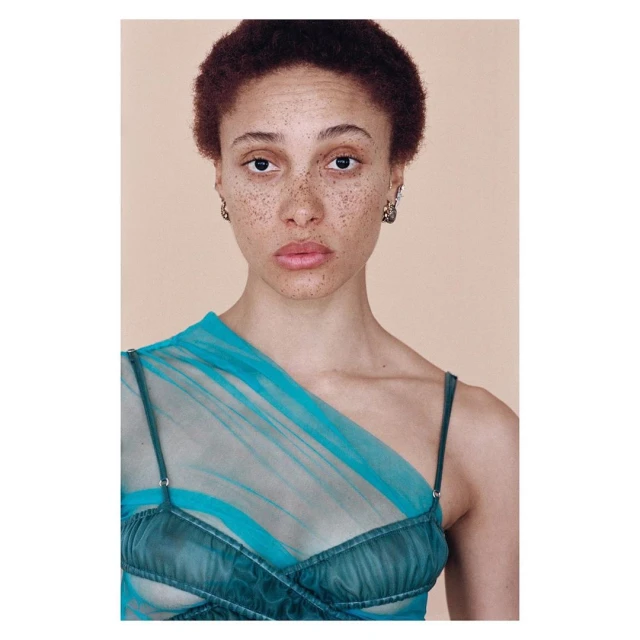 Portrait of a freckled adult female with short curly auburn hair, wearing a semi-sheer teal dress against a peach-colored backdrop.