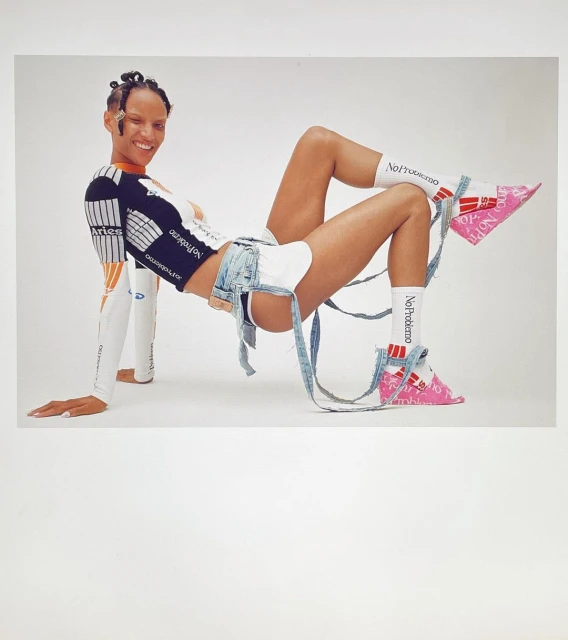 Smiling woman wearing a black and white long-sleeve shirt, denim shorts, pink shoes, and white socks posing in a studio.