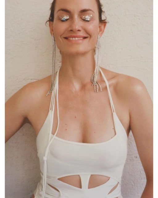 Adult woman smiles with eyes closed, wearing long, dangling crystal earrings and a white dress with cutout details, against a pale wall.