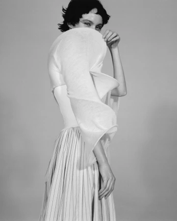 Monochrome portrait of a woman wearing a sculptural white top and pleated striped skirt against a plain grey background