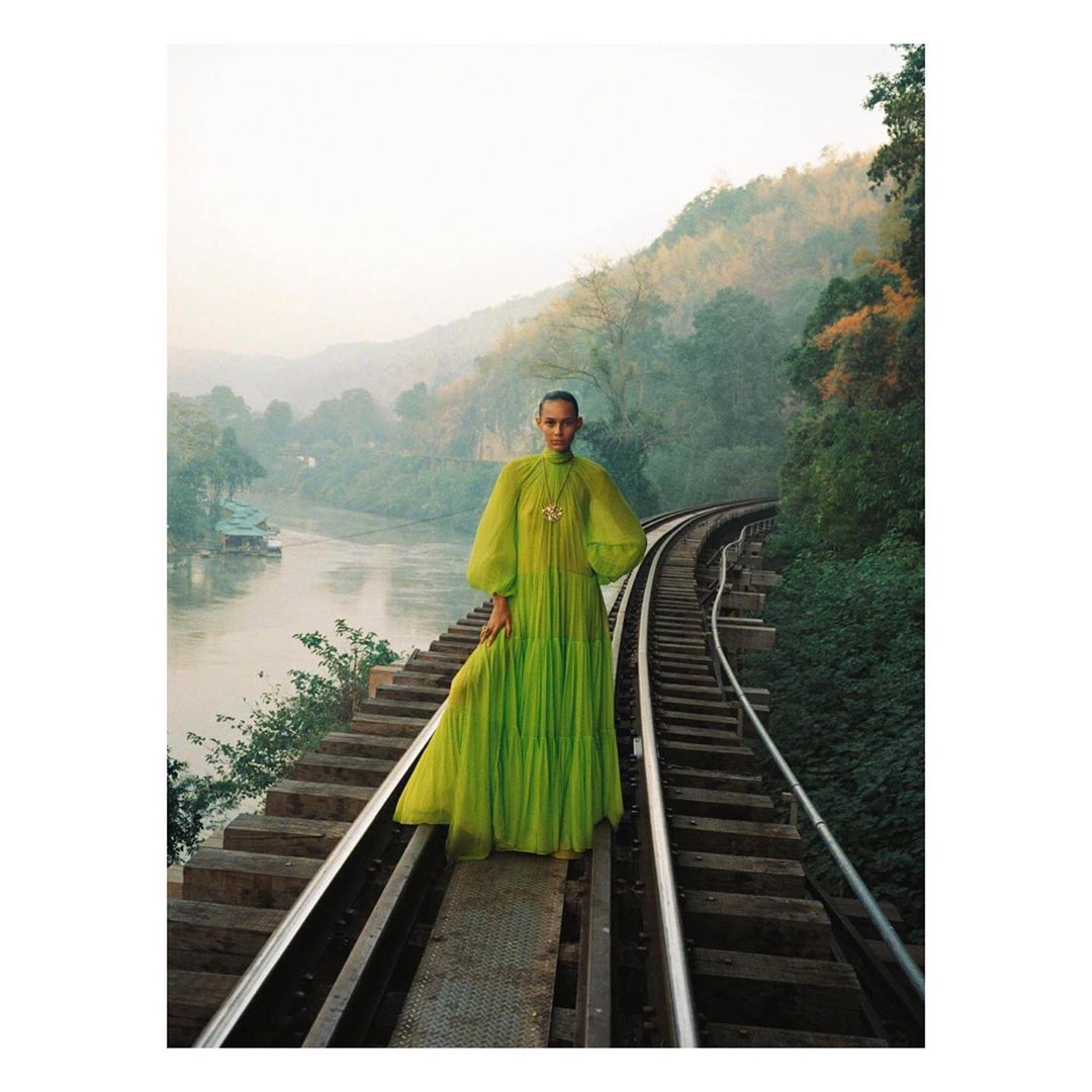 Woman in lime-green dress stands on train tracks overlooking river and mountains in a fashion editorial shot at eye-level.