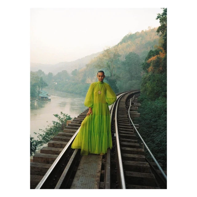 Woman in lime-green dress stands on train tracks overlooking river and mountains in a fashion editorial shot at eye-level.
