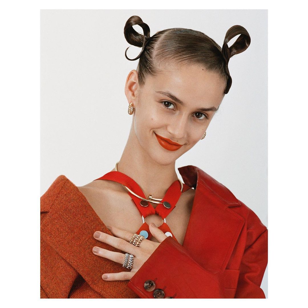 Portrait of an adult female model in a red jacket and gold jewelry with two buns against a white background in a studio.