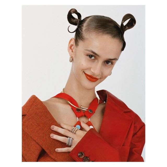 Portrait of an adult female model in a red jacket and gold jewelry with two buns against a white background in a studio.