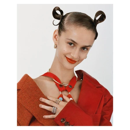 Portrait of an adult female model in a red jacket and gold jewelry with two buns against a white background in a studio.