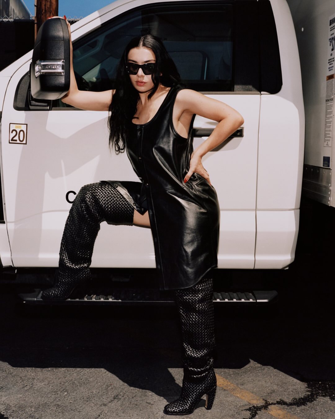 Adult female posing in a black dress and thigh-high boots leaning on a white truck under natural lighting, sunglasses and dark hair.
