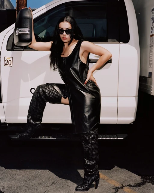 Adult female posing in a black dress and thigh-high boots leaning on a white truck under natural lighting, sunglasses and dark hair.