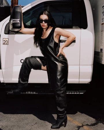 Adult female posing in a black dress and thigh-high boots leaning on a white truck under natural lighting, sunglasses and dark hair.