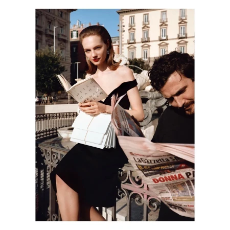 Elegant woman in black dress reading book by fountain, man reading newspaper in foreground, cityscape backdrop in outdoor setting.