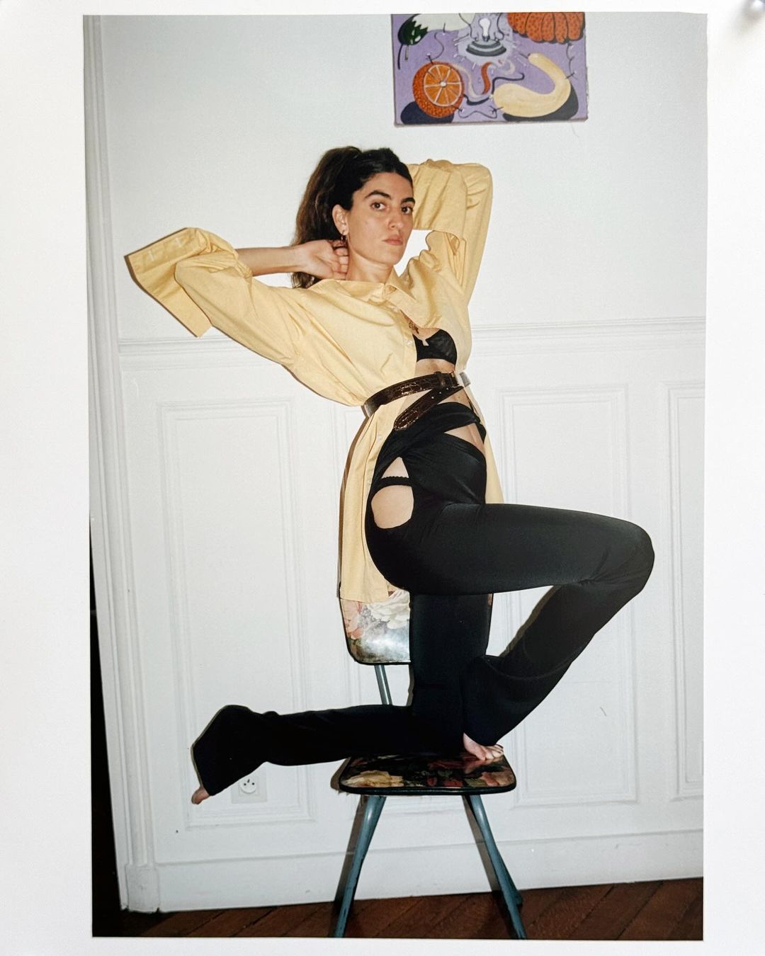A woman poses on a chair wearing an open yellow shirt and black cutout pants in a light indoor setting with a painting on the wall.