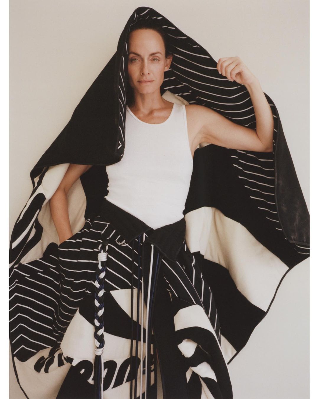 Adult woman wearing a white tank top and a large black and white patterned wrap or garment with a hood, posing in front of an off-white wall.