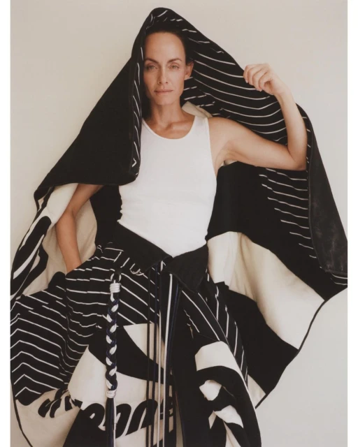Adult woman wearing a white tank top and a large black and white patterned wrap or garment with a hood, posing in front of an off-white wall.