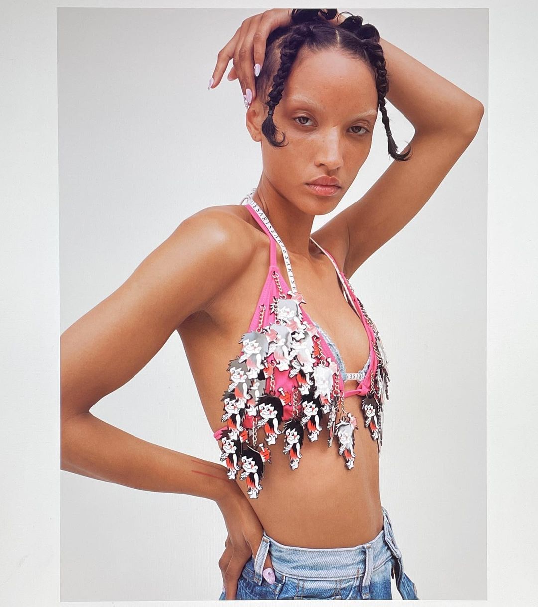 Fashion photo featuring a woman with braided hair posing in a pink halter top with cartoon character charms against a white background.