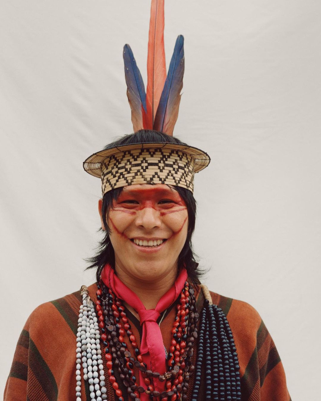 Smiling Indigenous man in traditional headdress with feathers and face paint, wearing beaded necklaces and a brown poncho against plain background.