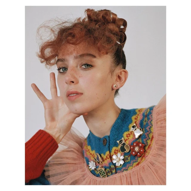 Young woman with curly red hair in a ruffled dress with pins poses in front of a white background in a studio shot.