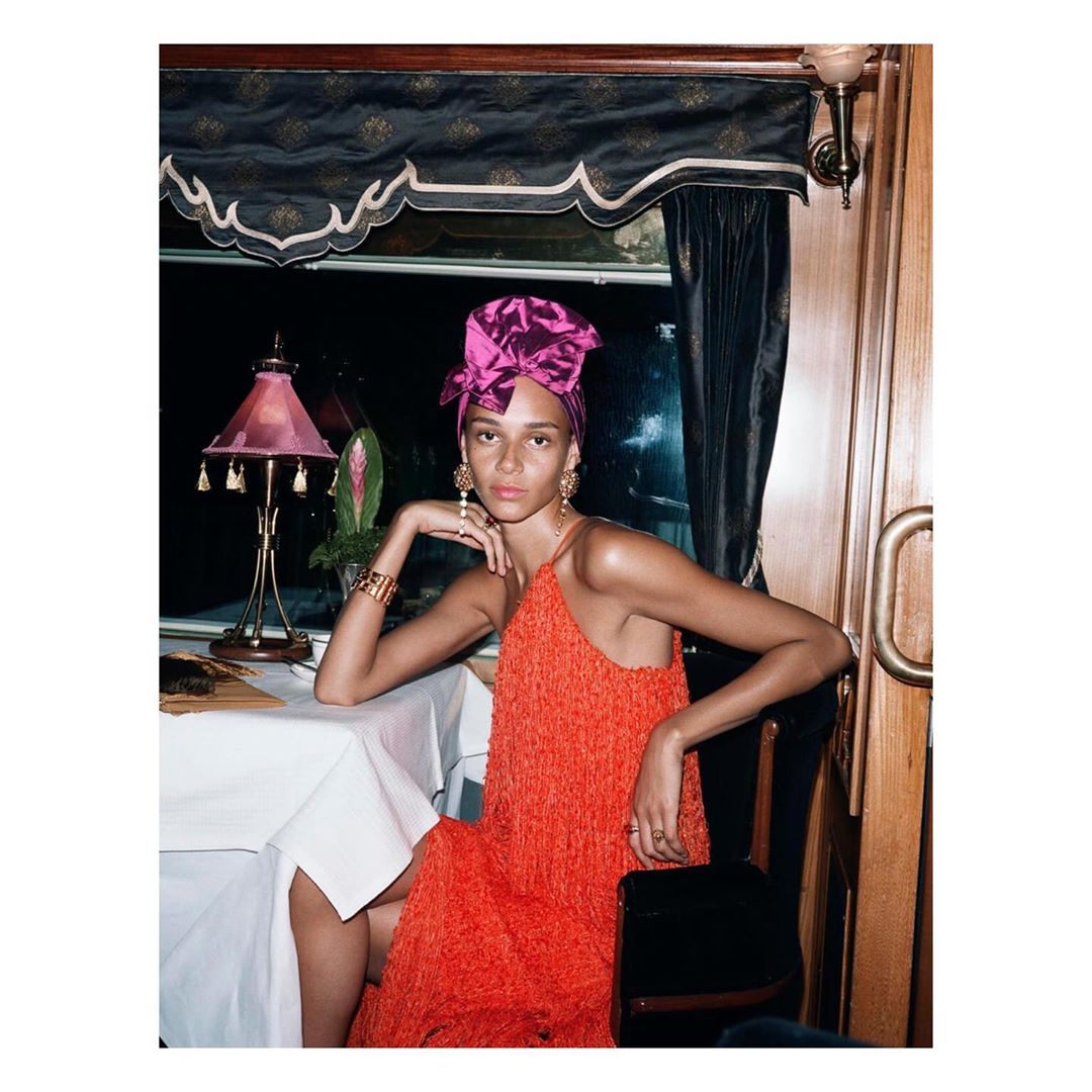 Elegant woman in orange fringe dress and purple turban seated at a table in a train dining car, accessorized with gold jewelry.