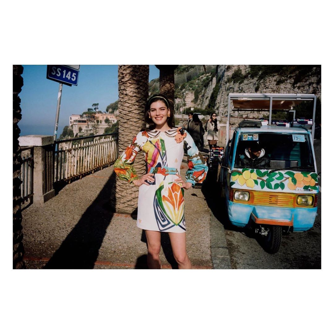 Woman in printed dress poses on Italian coast next to decorated cart with driver. Buildings and ocean stretch into distance under blue sky.