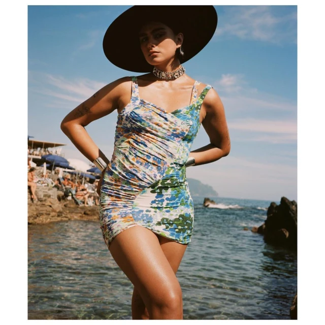 Woman wearing floral dress and large hat poses on rocky beach near ocean under blue sky, with sunbathers in background.