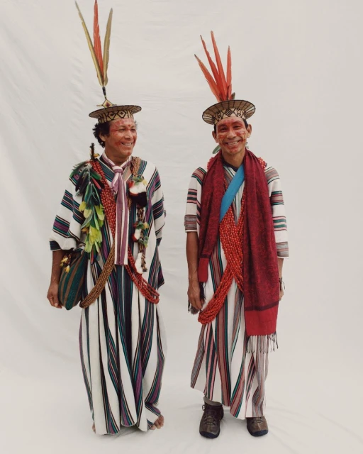 Two Indigenous men in traditional striped clothing, headwear with feathers, and adornments, standing indoors against a plain white backdrop.