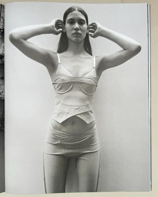 Monochrome portrait of a woman posing in a minimalist outfit, hands near her head, against a simple background, emphasizing clean fashion.
