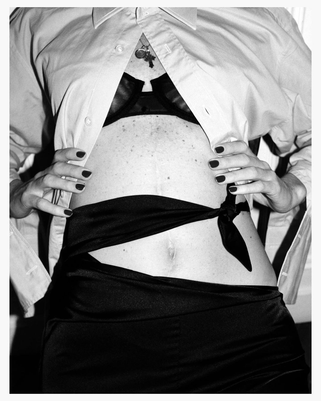 Monochrome image of a torso with an open white shirt revealing a black bra, accentuated by a knotted black sash, and polished nails.
