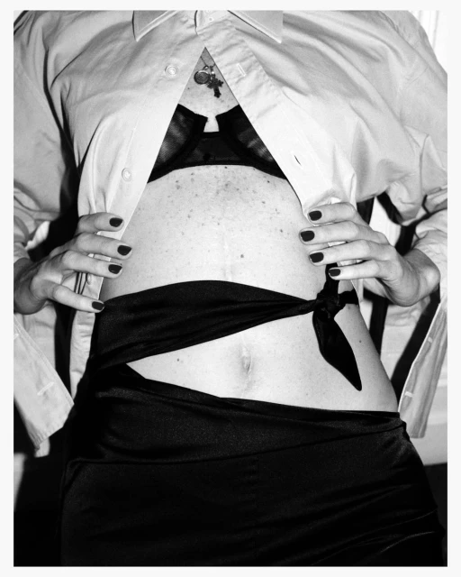 Monochrome image of a torso with an open white shirt revealing a black bra, accentuated by a knotted black sash, and polished nails.