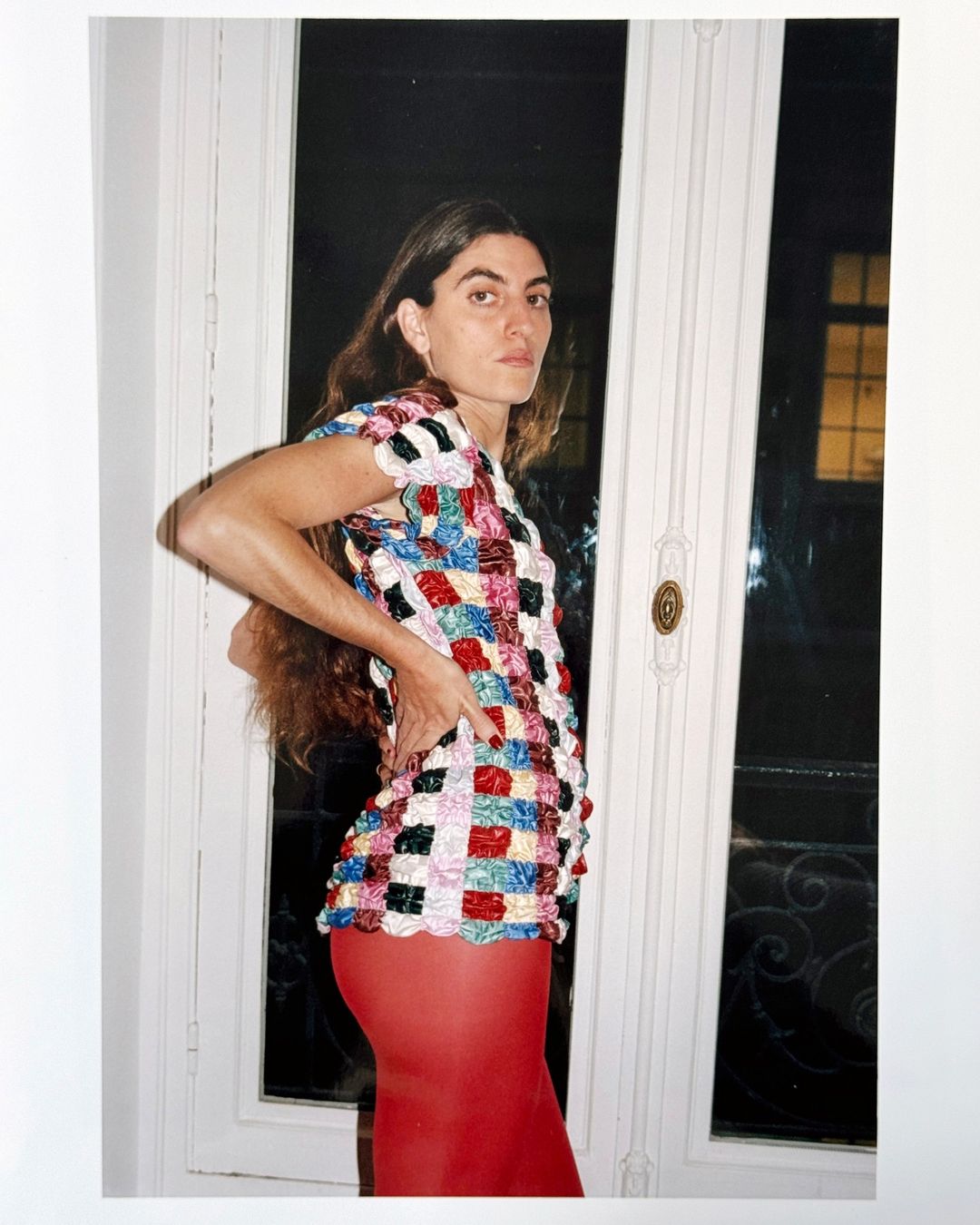 Woman poses indoors near a window, wearing a multicolored patterned top and red leggings, capturing a candid and fashion-forward aesthetic.
