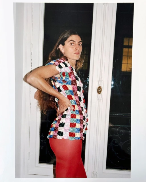 Woman poses indoors near a window, wearing a multicolored patterned top and red leggings, capturing a candid and fashion-forward aesthetic.